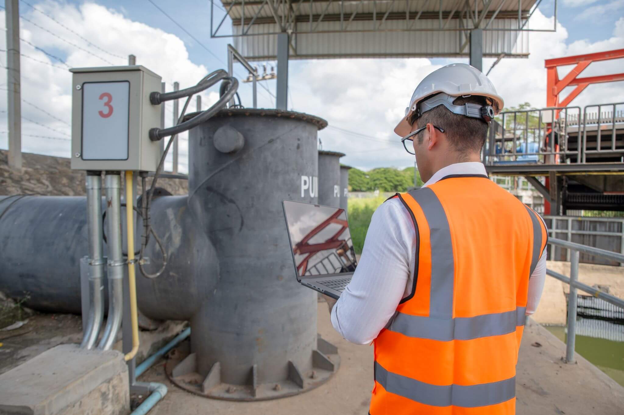 Engineer in safety vest and hard hat using a laptop to inspect industrial equipment and a control box