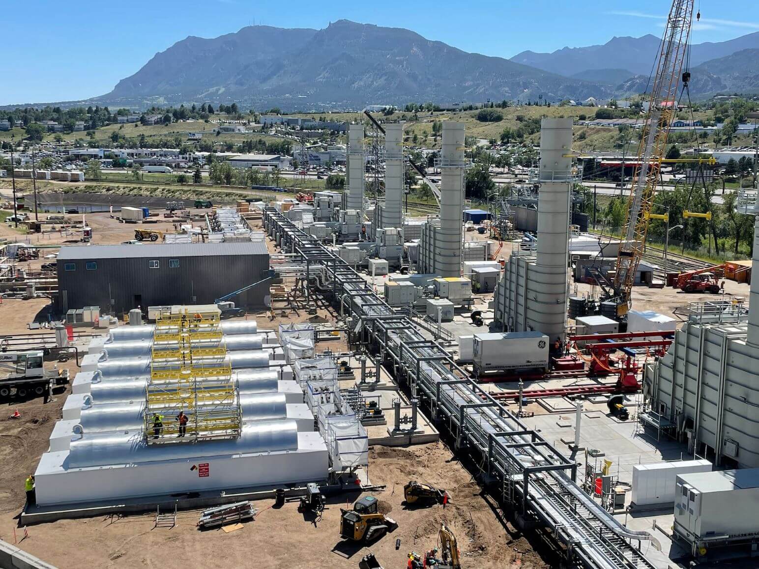 Utility-scale gas turbine power plant under construction with exhaust stacks, pipe racks and balance-of-plant systems against a mountain backdrop